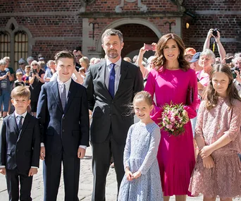 A family poses outside a historic building, with the woman in pink holding flowers, surrounded by children in formal attire.