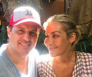 A man in a baseball cap and a woman with pulled-back hair smiling at each other outdoors.
