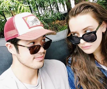 Two individuals wearing sunglasses sitting together, engaged in eye contact with a backdrop of green plants.