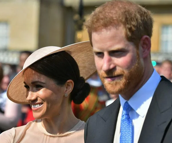 A couple smiling at an outdoor event, with the woman wearing a hat and the man in a suit with a blue tie.