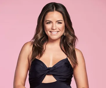 A woman with long brown hair in a black dress smiles against a pink background.
