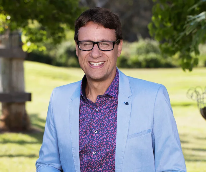 Man in glasses and a blue blazer smiling outdoors with trees and grassy background.