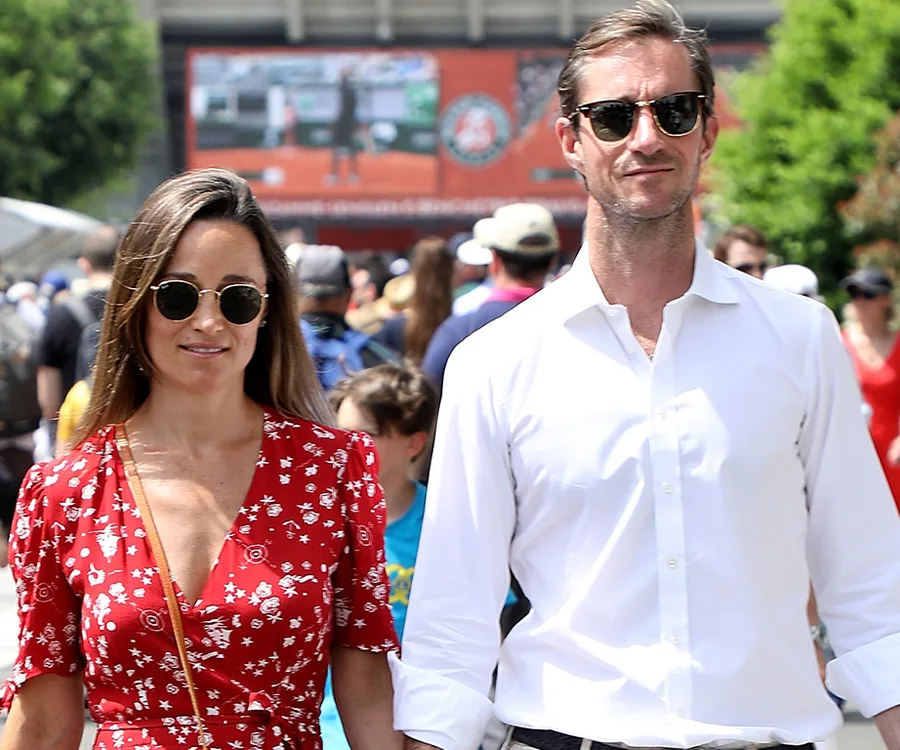 A woman in a red dress and a man in a white shirt walking outdoors, with a crowd in the background at an event.