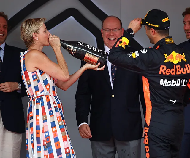 "Princess Charlene of Monaco drinks champagne at the Grand Prix podium celebration, with racing driver and guests."