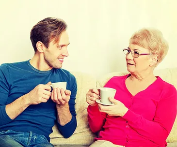 A man and an older woman sitting on a couch, holding mugs and engaging in a conversation.