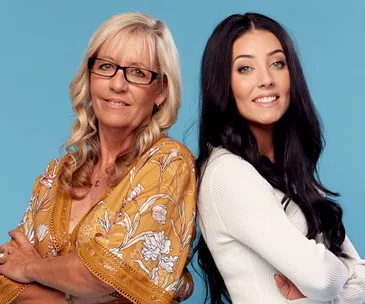 Two women standing back-to-back, one with blonde hair wearing glasses, and the other with long black hair, smiling.