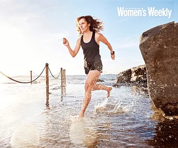 Woman running through water, wearing athletic gear, smiling with determination. Ocean and rocks in the background, bright daylight.