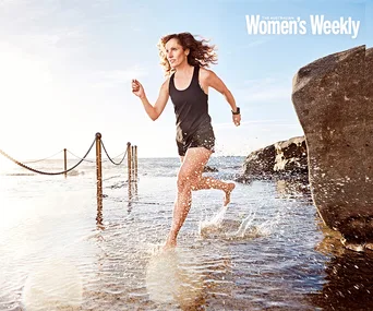 Woman running through water, wearing athletic gear, smiling with determination. Ocean and rocks in the background, bright daylight.