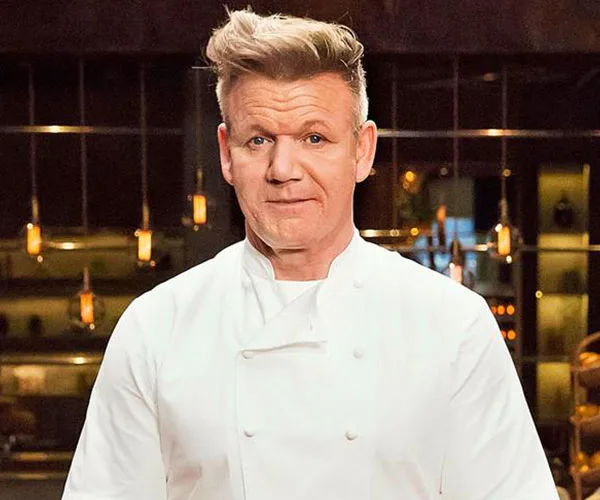 Chef in a white uniform stands in a kitchen setting with dim lighting and hanging lights in the background.