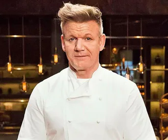 Chef in a white uniform stands in a kitchen setting with dim lighting and hanging lights in the background.