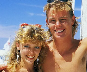 Two people smiling at the camera in a sunny setting, with one of them wearing sunglasses on their head.