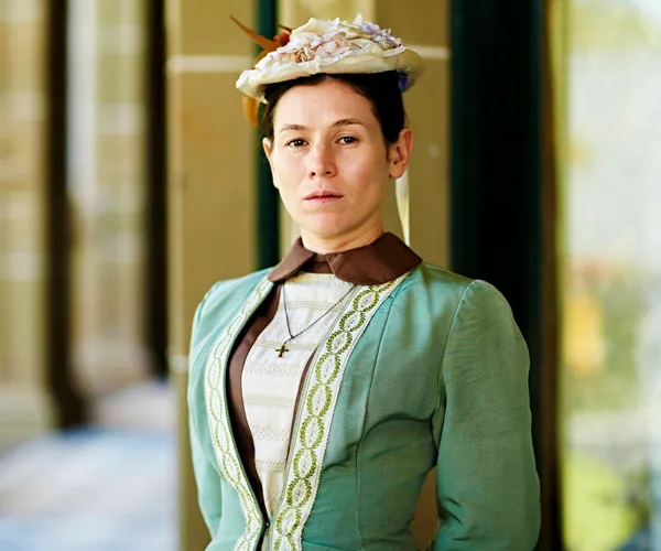 A woman in period costume with a hat, from "Picnic at Hanging Rock," stands outside a building.