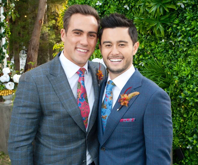 Two men in suits smiling at a wedding with a leafy background.