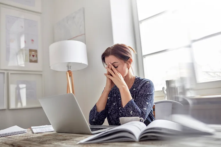 Stressed woman at a desk with a laptop, hands on face, in a bright office setting.