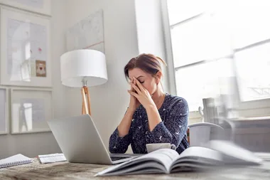 Stressed woman at a desk with a laptop, hands on face, in a bright office setting.