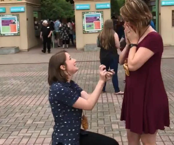 Person kneeling and proposing with ring while other gasps, surprised and emotional, in public area with onlookers.