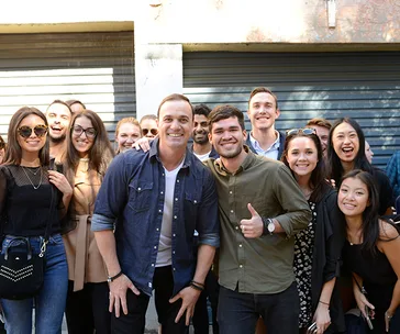 Group of people smiling, with one person giving a thumbs up, standing outside in front of a building with a shutter.