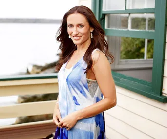 Female standing outdoors near a window, wearing a blue and white dress, smiling. Background shows water and trees.