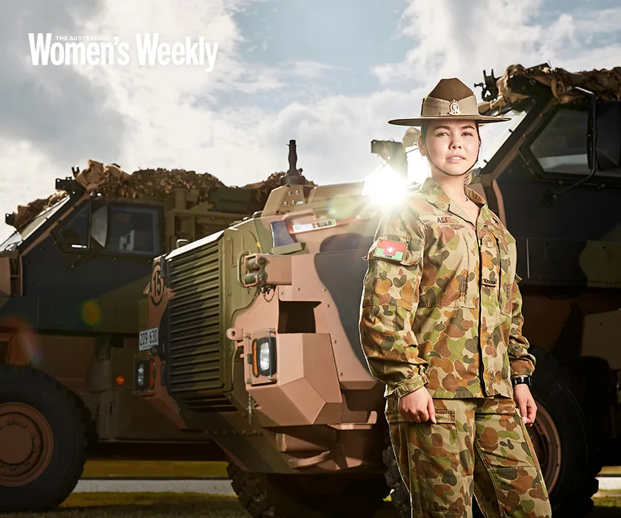 Australian Army soldier in uniform stands confidently next to an armored vehicle under a partly cloudy sky.