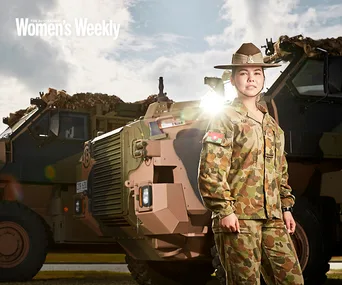 Australian Army soldier in uniform stands confidently next to an armored vehicle under a partly cloudy sky.