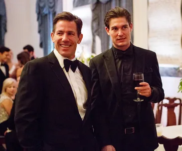 Two men in tuxedos at a formal event, one holding a wine glass, smiling and conversing.