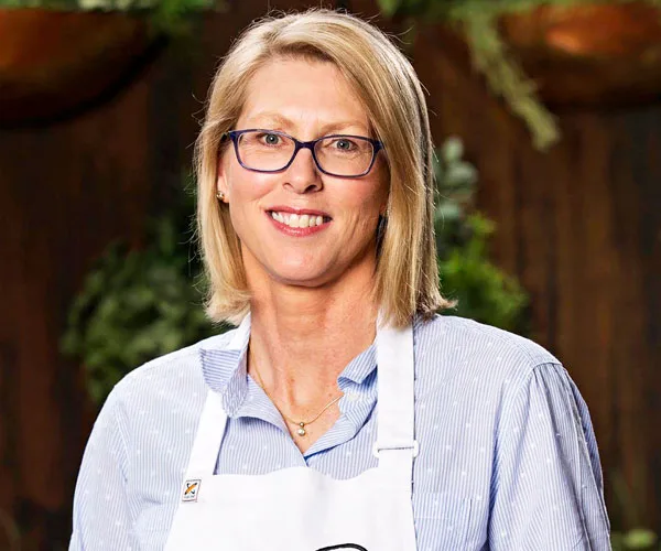 Genene Dwyer from MasterChef Australia wearing glasses and a chef apron, smiling at the camera.