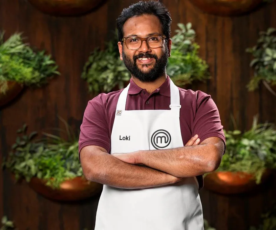 Man wearing glasses and a "Loki" MasterChef apron, standing with arms crossed in front of a plant-decorated wall.