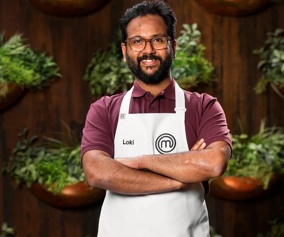Man wearing glasses and a "Loki" MasterChef apron, standing with arms crossed in front of a plant-decorated wall.