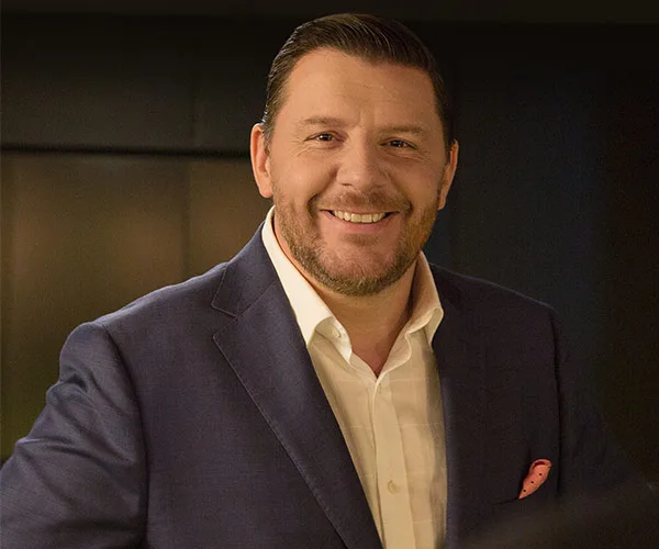 Smiling man in a blue suit and white shirt, standing against a dark background.