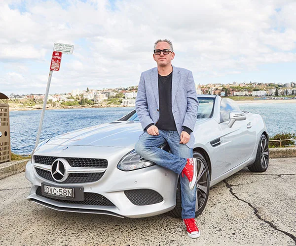 Person in sunglasses leans on a silver Mercedes by the seaside, wearing a light blazer, jeans, and red sneakers.