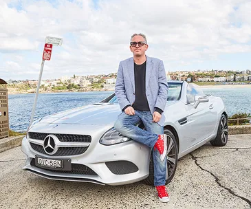 Person in sunglasses leans on a silver Mercedes by the seaside, wearing a light blazer, jeans, and red sneakers.