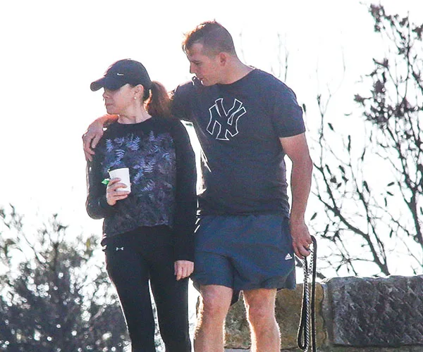 A couple outdoors, man with arm around woman holding a cup, both in casual sportswear, trees in background.