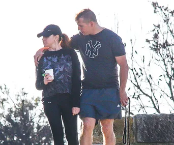 A couple outdoors, man with arm around woman holding a cup, both in casual sportswear, trees in background.