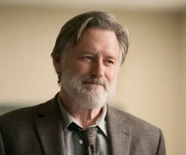 Man with gray hair and beard in a suit, smiling softly; scene from "The Sinner" season 2.