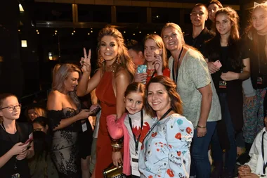 Group of fans posing with a woman in an orange dress flashing a peace sign, with a crowd behind them.