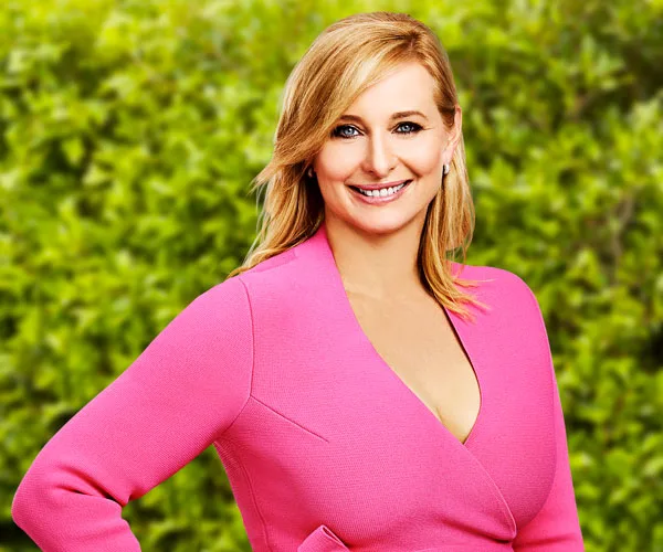 A woman with blonde hair wearing a pink outfit smiles in front of a green leafy background.