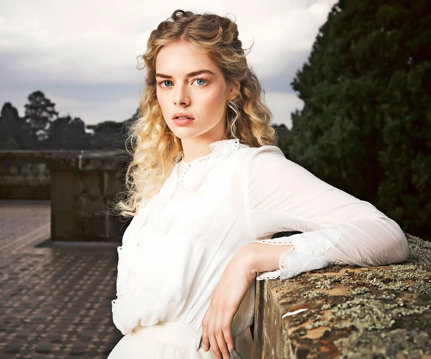 A young woman with curly blonde hair in a white dress stands outdoors, leaning on a stone wall with trees in the background.