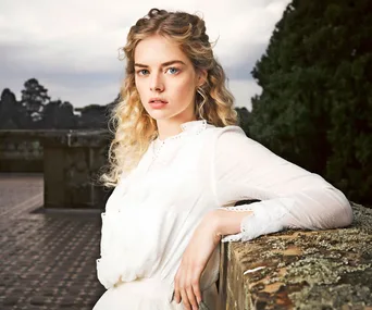 A young woman with curly blonde hair in a white dress stands outdoors, leaning on a stone wall with trees in the background.
