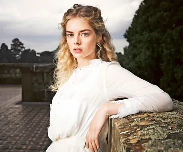 A young woman with curly blonde hair in a white dress stands outdoors, leaning on a stone wall with trees in the background.