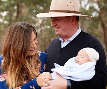 A couple smiles at each other while holding a baby wrapped in a blue blanket outdoors.