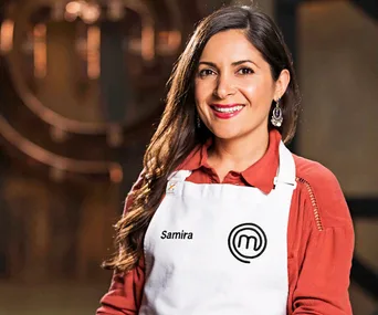 "Woman in a red shirt wearing a white MasterChef apron with 'Samira' printed on it, smiling in a kitchen setting."