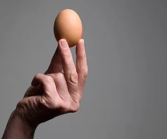 A hand holding a brown egg between the fingers against a gray background.