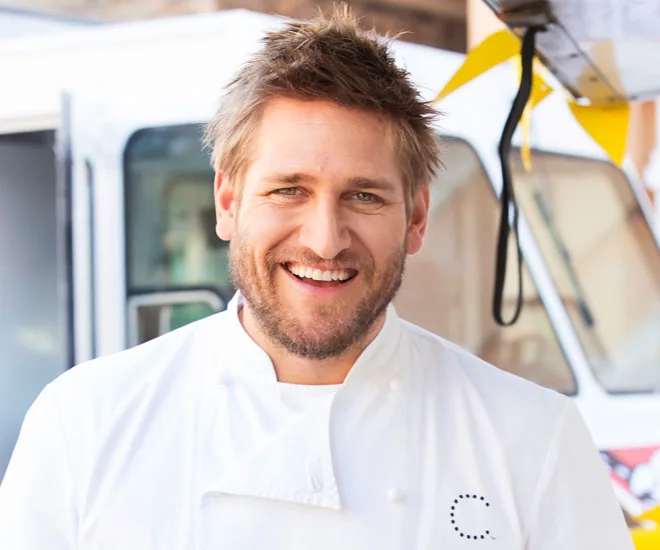 Chef in a white coat smiling outdoors near a food truck.