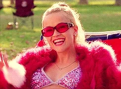 Elle Woods from "Legally Blonde," wearing pink sunglasses, a sparkly top, and a fur coat, smiles and waves outdoors.