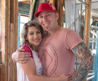 Man and woman smiling, embracing in a partially constructed house. Man wears a red cap, woman has curly hair and red lipstick.