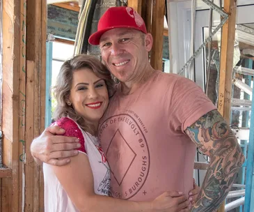 Man and woman smiling, embracing in a partially constructed house. Man wears a red cap, woman has curly hair and red lipstick.