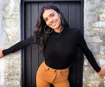 A woman in a black top and brown pants smiles with arms outstretched against a rustic, textured wall.