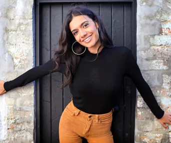 A woman in a black top and brown pants smiles with arms outstretched against a rustic, textured wall.