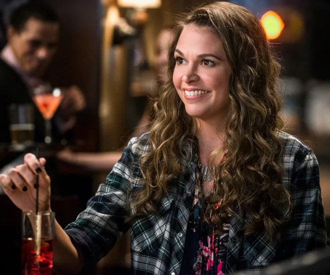 A woman with wavy hair smiles at a bar, holding a red cocktail with a straw, wearing a plaid shirt.