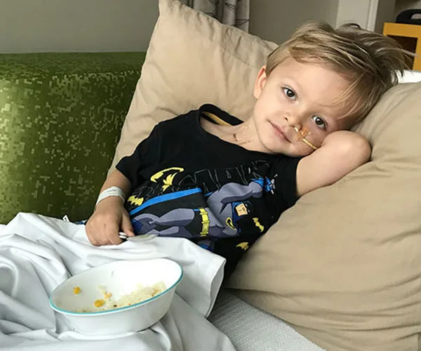 Young boy with a nasal tube, wearing a Batman shirt, reclines in bed with a bowl of food beside him.